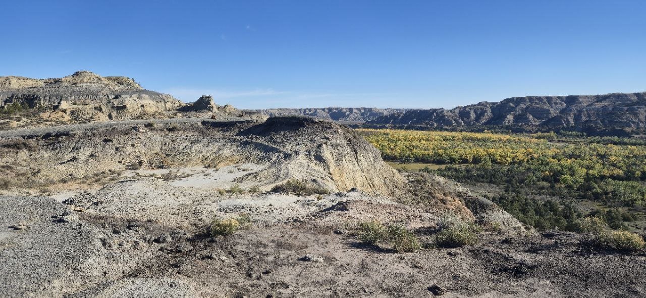 A view across the badlands toward the river bottom, with the trail&rsquo;s rocky terrain in the foreground.