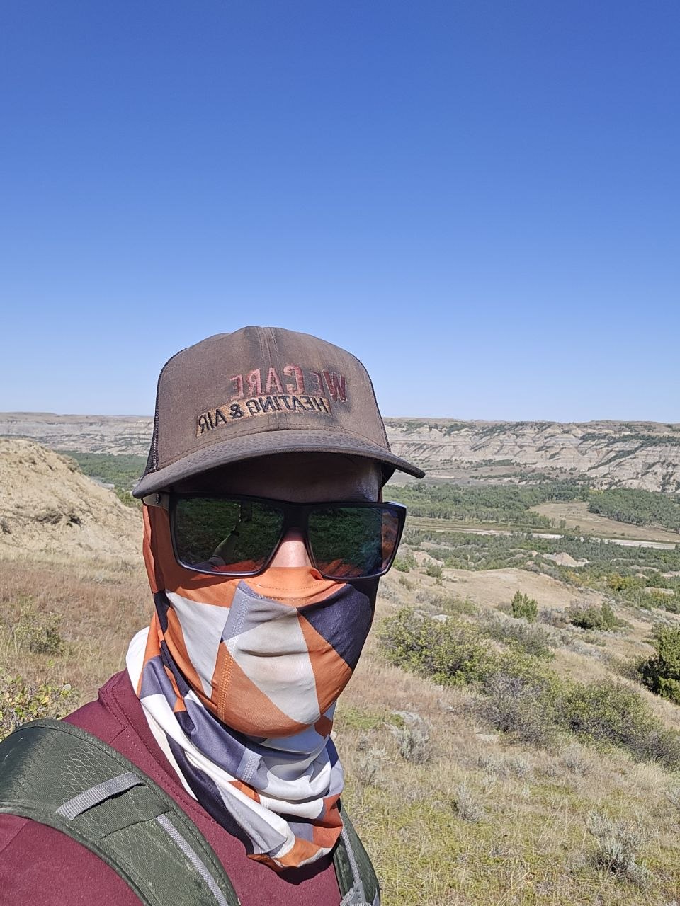 A trail selfie above the badlands valley, wearing sunglasses, a ballcap, and a PackLite Life geometric neck gaiter.