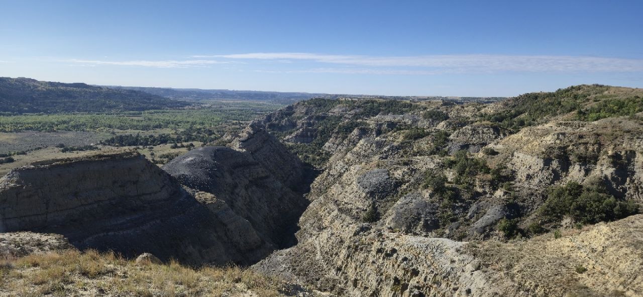 A sweeping badlands view from Sperati Point, with the Little Missouri River winding through a green cottonwood corridor below.