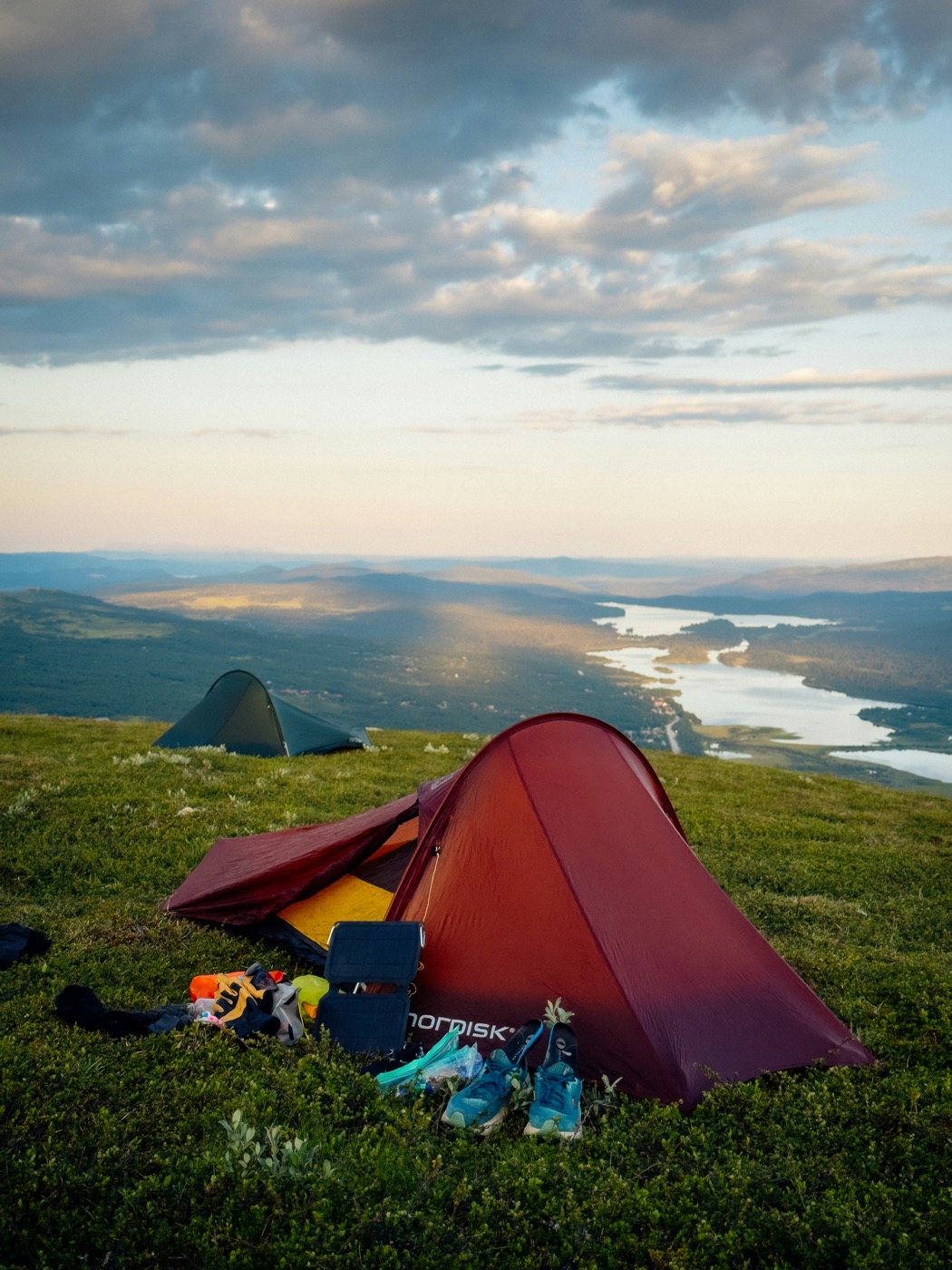 A deep red Nordisk tunnel tent pitched on a grassy hilltop overlooking a lake at golden hour