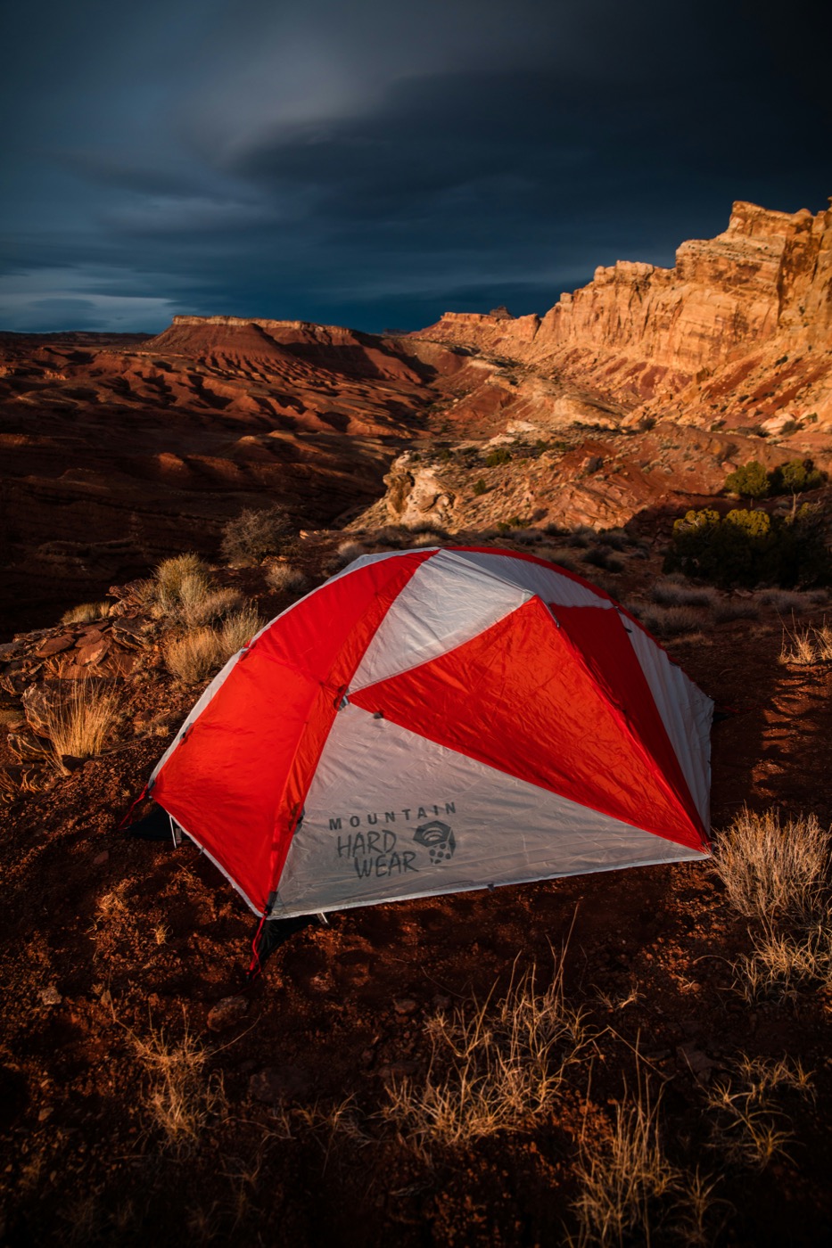 A red and white Mountain Hardwear geodesic tent pitched in a dramatic desert canyon at dusk